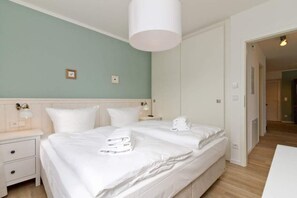 Quarto de hotel com duas camas, uma mesa de cabeceira branca, uma luminária na parede e uma porta que dá para outro cômodo.