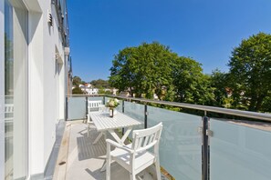 A balcony with a white table and chairs, a glass railing, and a view of trees and buildings.