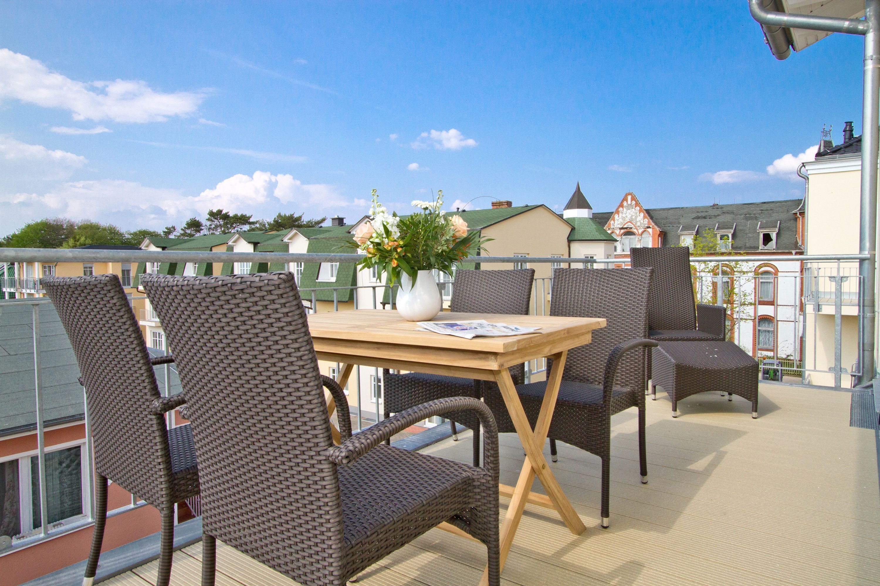 A balcony with wicker chairs and a table, a vase with flowers, and a view of buildings in the background.