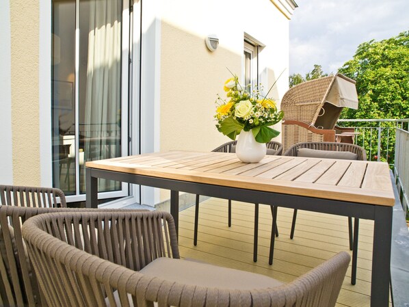 Eine Terrasse im Freien mit Holztisch und Korbstühlen, einem Blumenstrauß und Blick auf ein Gebäude mit großen Fenstern.