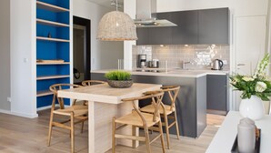 A modern kitchen with a central island, wooden chairs, a hanging light fixture, and a blue shelving unit. - Dünenpalais, App. 10 (Heringsdorf)