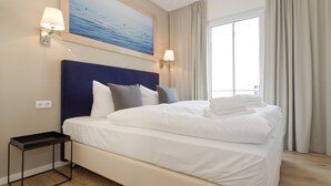 A hotel room with a large bed, two bedside lamps, a painting above the bed, a nightstand, and a window with curtains. - Dünenpalais, App. 10 (Heringsdorf)