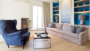A modern living room with a blue armchair, a beige sofa, and a coffee table. There is a fireplace, a large window with a view of the sea, and blue shelving with decorative items. - Dünenpalais, App. 10 (Heringsdorf)