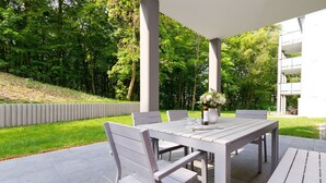 Eine Terrasse im Freien mit einem weißen Tisch und Stühlen, umgeben von üppiger, grüner Natur und einem modernen Gebäude im Hintergrund.