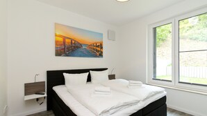 Ein modernes Schlafzimmer mit einem großen Bett, einem Nachttisch, einer Wandleuchte und einem Fenster mit Blick auf Grünflächen.