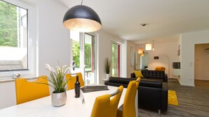 Ein modernes Wohnzimmer mit einer schwarzen Couch, gelben Sesseln und einem Esstisch, auf dem ein Topfpflanze und eine Weinflasche stehen.