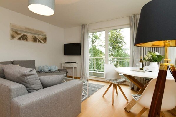 A modern living room with a grey sofa, a dining table with chairs, a TV mounted on the wall, and a balcony with a view of greenery.