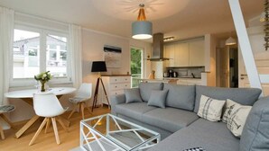 A modern living room with a grey sofa, a glass coffee table, and a dining area with white chairs and a small table. - Villa Barbara, App. 09 (Heringsdorf)