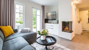 A modern living room with a grey sofa, a coffee table with a vase and magazine, a fireplace, and a TV mounted on the wall. - Ostseedomizil Bansin, App. 1.1 (Heringsdorf)