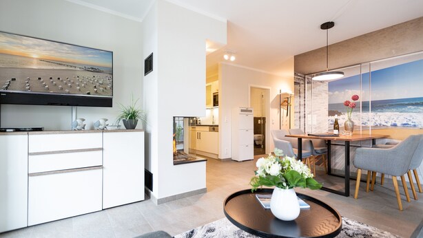 A modern living room with a flat-screen TV, a dining area with a wooden table and chairs, and a kitchen in the background. - Ostseedomizil Bansin, App. 1.1 (Heringsdorf)