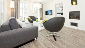 Una sala de estar moderna con un sofá gris, una silla negra y una mesa de centro blanca. Hay dos ventanas con cortinas, un televisor de pantalla plana montado en la pared y un estante con objetos decorativos.