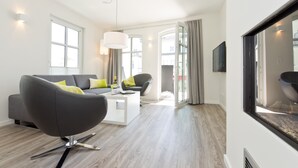 Una sala de estar moderna con un sofá gris, una mesa de centro y una silla. Se ve un ventanal con cortinas, un televisor de pantalla plana montado en la pared y un candelabro.