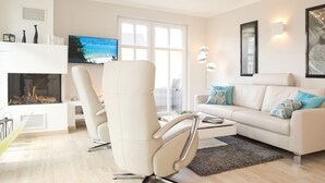 Una sala de estar moderna con un sofá blanco, dos sillones blancos, una chimenea, un televisor de pantalla plana y un ventanal grande.