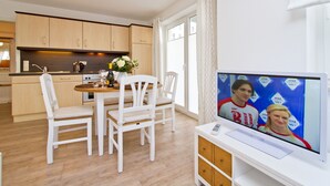 A modern kitchen with wooden cabinets, a dining area with white chairs, and a television displaying a Visa advertisement. - Apartment house Neuer Weg, App. 02 (Heringsdorf)