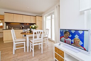 A modern kitchen with wooden cabinets, a dining area with white chairs, and a television displaying a Visa advertisement. - Apartment house Neuer Weg, App. 02 (Heringsdorf)