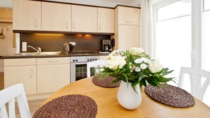 A modern kitchen with wooden cabinets, a stove, and a coffee maker. There is a dining table with a vase of flowers and woven placemats. - Apartment house Neuer Weg, App. 02 (Heringsdorf)