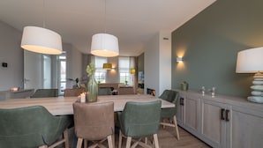 A modern dining area with a wooden table and green upholstered chairs, pendant lights, and a cabinet with decorative items. - Villa in Zeeland near Clean Beaches (Colijnsplaat)
