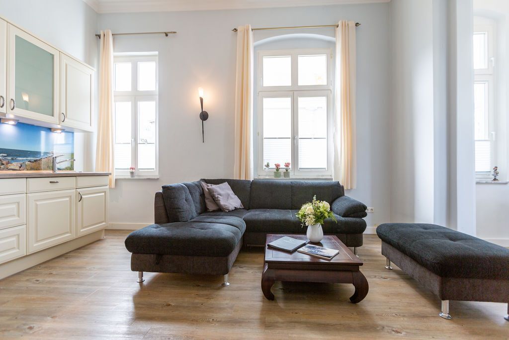 Un salon avec un canapé d’angle gris foncé, une table basse en bois et une petite table d’appoint sur laquelle est posé un vase de fleurs. Une grande fenêtre, ornée de rideaux, laisse entrer la lumière, et un luminaire mural éclaire la pièce. La fenêtre offre une vue sur la mer.