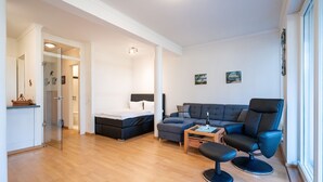 A modern living room with a sofa, a coffee table, and a chair. There is a bed with a headboard and a small table beside it. The room has wooden flooring and white walls. - Villa Marfa, App. 02 (Heringsdorf)