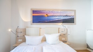A double bed with white bedding, a wooden headboard, and a wall-mounted painting of a pier at sunset. - Villa Limora, App. 02 (Heringsdorf)