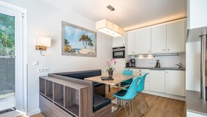 A modern kitchen with white cabinets, a dining area with a wooden table and blue chairs, and a painting of a beach scene on the wall. - Villa Limora, App. 02 (Heringsdorf)
