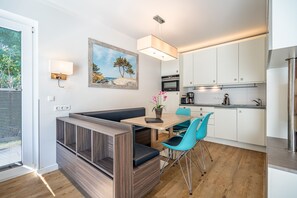 Une cuisine moderne avec des meubles blancs, un coin repas avec une table en bois et des chaises bleues, et un tableau représentant une scène de plage accroché au mur.