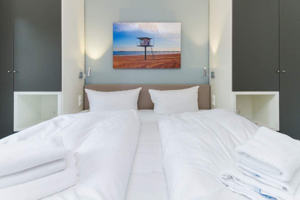 A hotel room with a bed, two bedside tables, a painting of a lifeguard tower, and grey wardrobes.