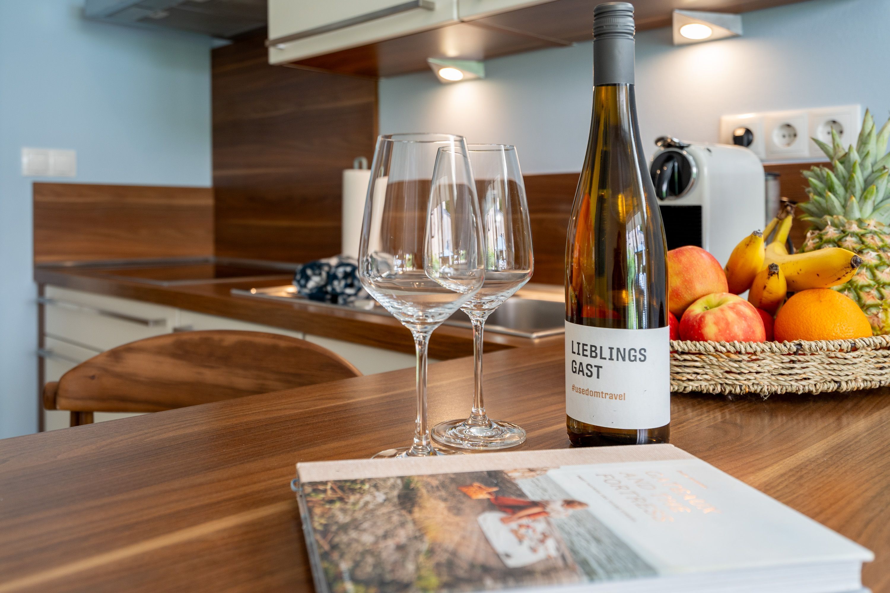 A kitchen with a wooden table, two wine glasses, a bottle of wine, and a basket of fruit.