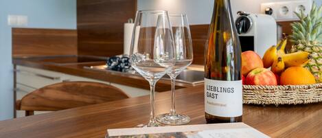 A kitchen with a wooden table, two wine glasses, a bottle of wine, and a basket of fruit.