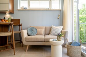 Ein Wohnzimmer mit einem beige farbenen Sofa, einem Holztisch, einem Korbwäschesessel und einem Fenster mit Blick auf Grünflächen.