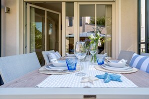 Una mesa de comedor dispuesta con cristalería azul, platos blancos y decoraciones de estrellas de mar, con vistas a un edificio a través de una puerta de vidrio.