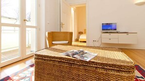 Une pièce avec un pouf en osier, un téléviseur posé sur un meuble blanc et une porte donnant sur une autre pièce.