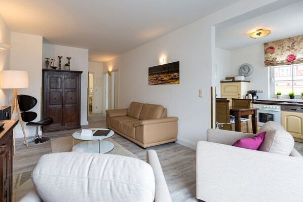 Un salon moderne avec un canapé beige, un canapé blanc et une table basse ronde. On y trouve également un meuble en bois et une chaise noire. On aperçoit à l’arrière-plan un coin cuisine avec une cuisinière et des placards.