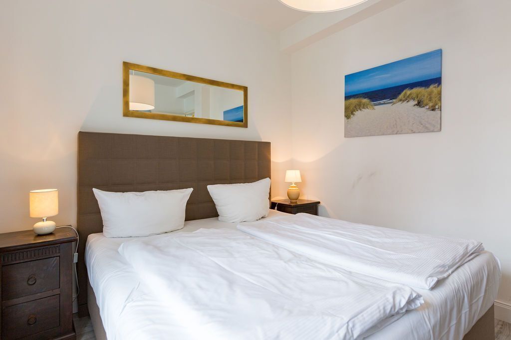 Una habitació d'hotel amb un llit gran, una tauleta de nit amb una làmpada, un mirall i un quadre amb tema de platja a la paret.