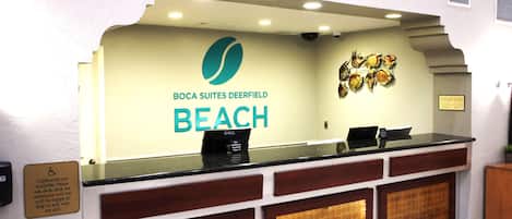 Un mostrador de recepción con un cartel de Boca Suites Deerfield Beach, una computadora Dell y un anuncio sobre archivadores disponibles.