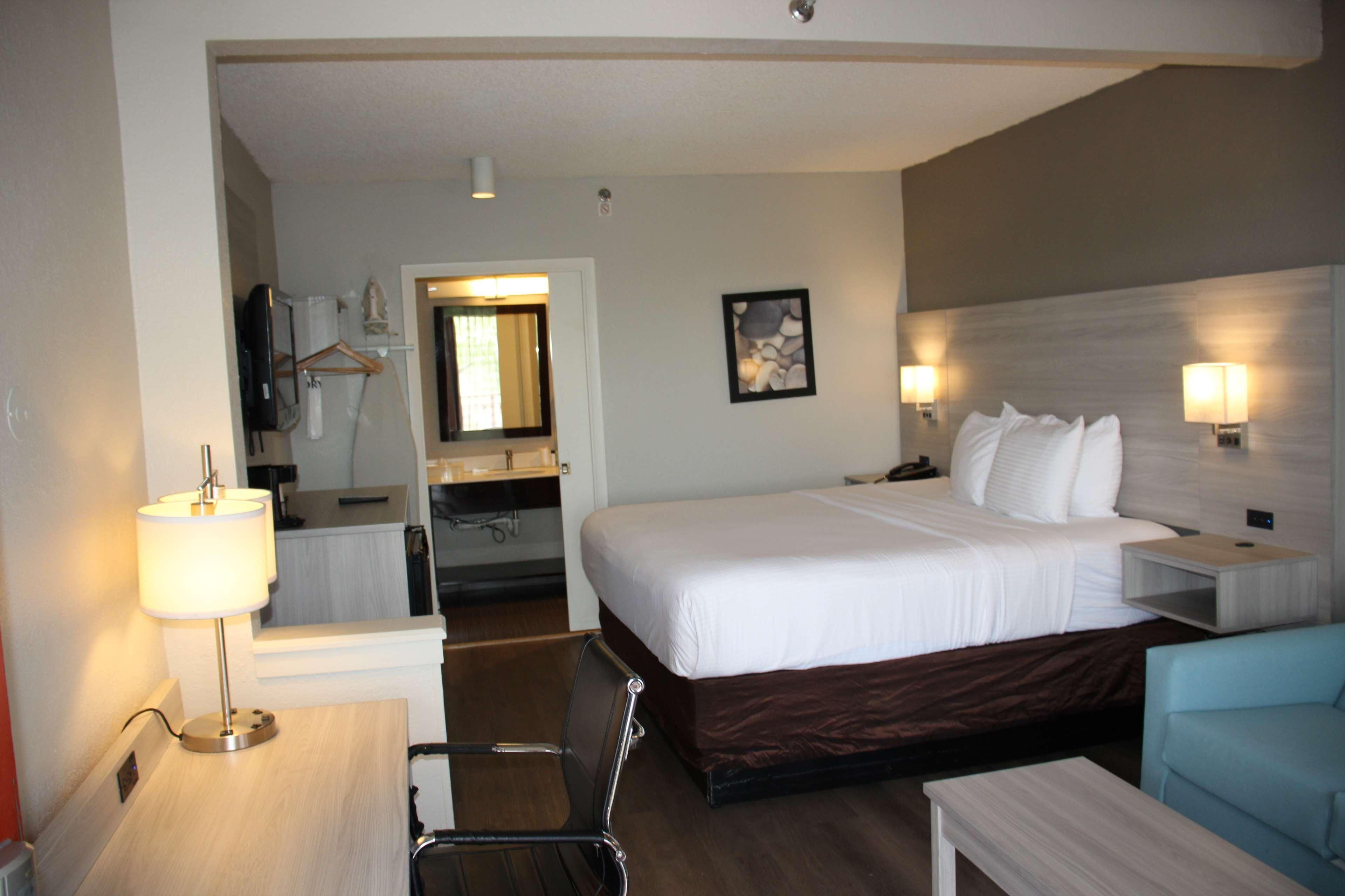 Une chambre d’hôtel comprenant un lit, des tables de chevet, un bureau, une chaise, un miroir, une salle de bain et un tableau accroché au mur.