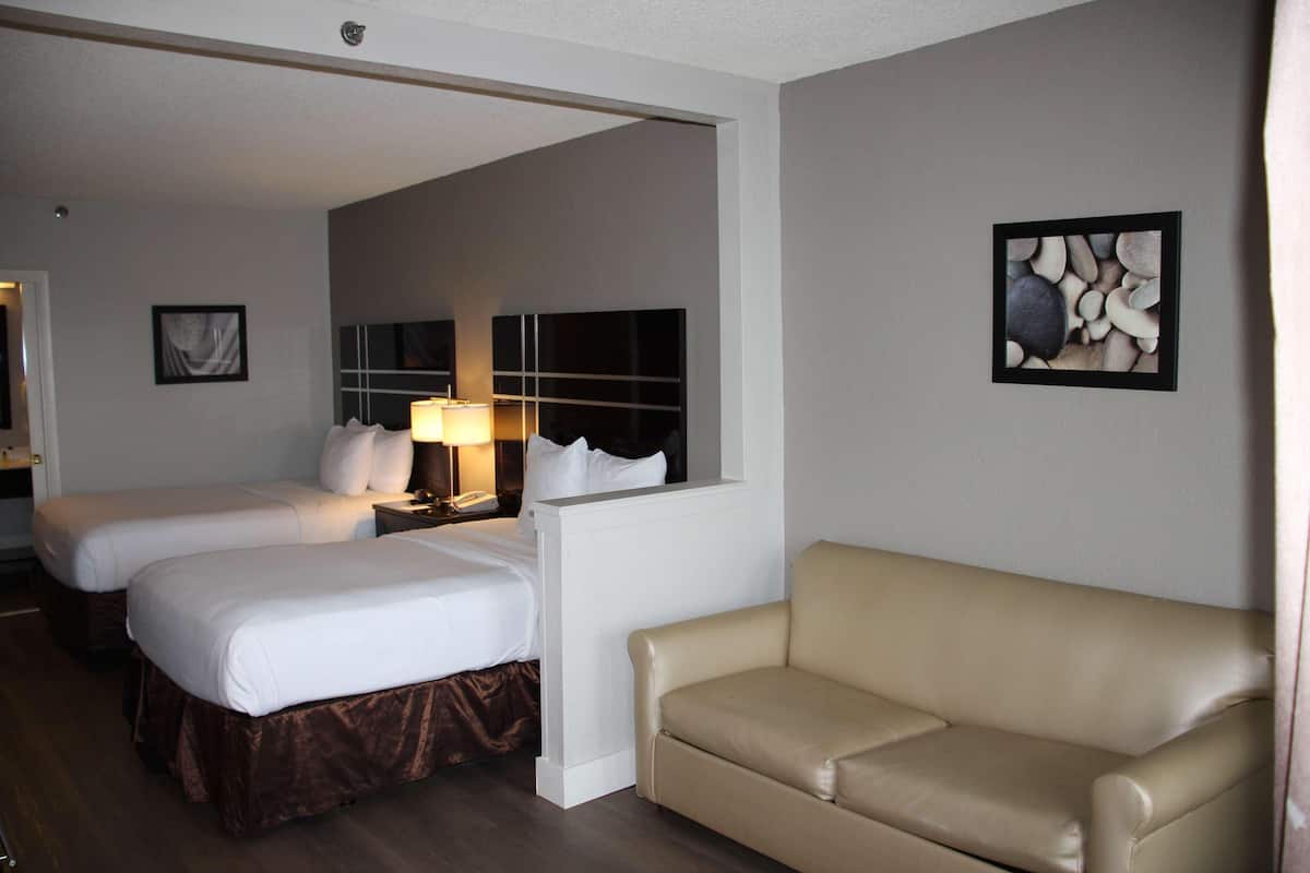 Una camera d'albergo con due letti, un divano beige, un comodino con una lampada e un quadro incorniciato appeso al muro.