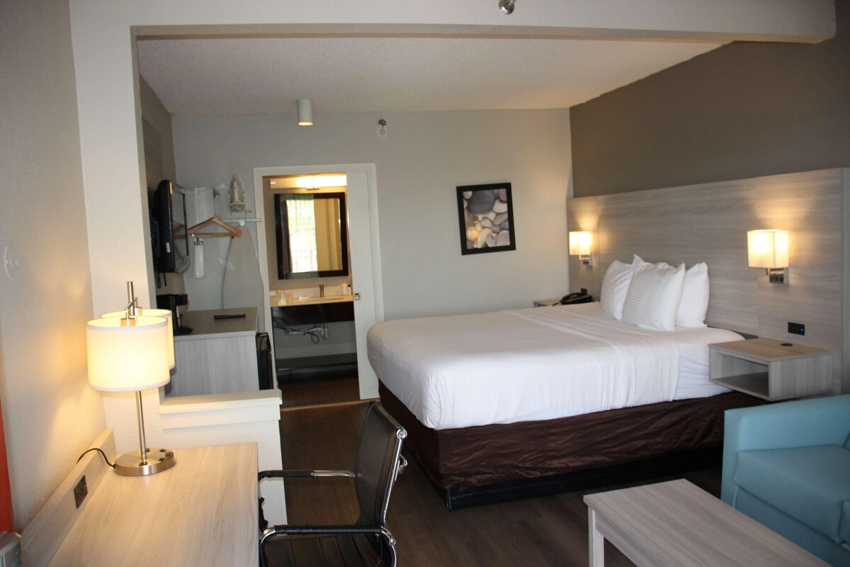A hotel room with a bed, bedside tables, a desk, a chair, a mirror, a bathroom, and a painting on the wall.
