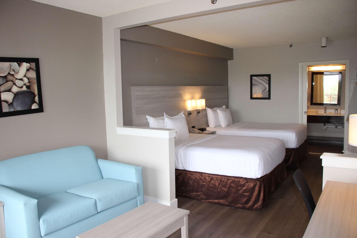 A hotel room with two beds, a light blue sofa, a small table, and a bathroom visible through an open door.