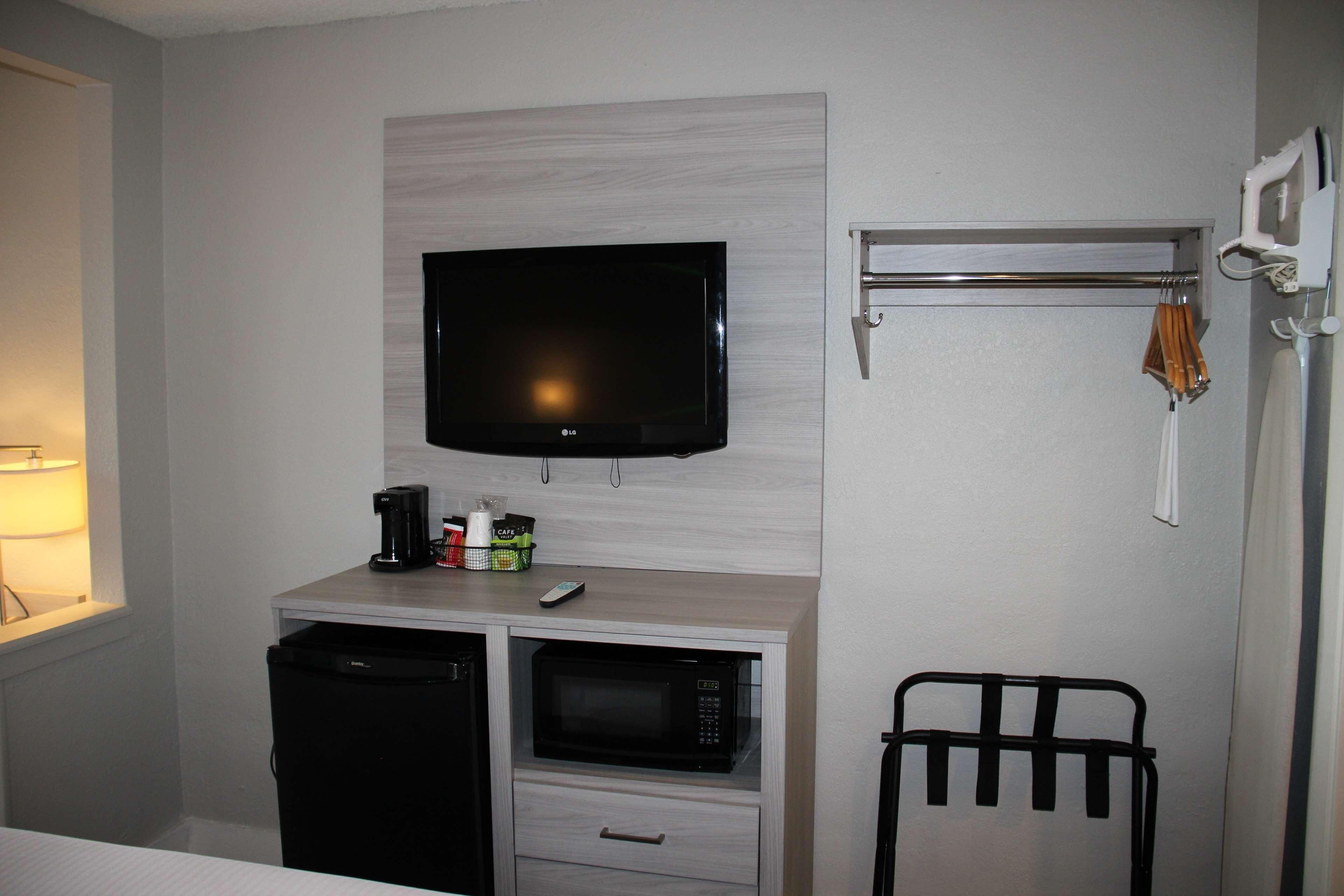 Ein kleines Hotelzimmer mit Flachbildfernseher an der Wand, einer Mikrowelle, einem kleinen Kühlschrank und einem Metallregal mit einem Handtuch.