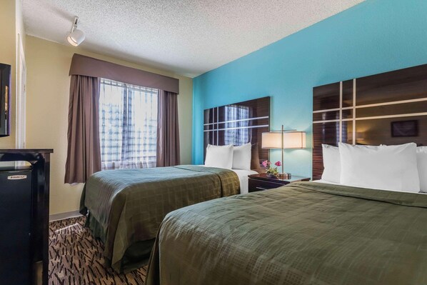 Quarto de hotel com duas camas, uma escrivaninha, uma luminária e uma janela grande com cortinas.
