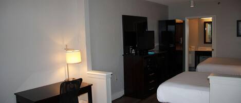 Habitación de hotel con escritorio, silla, televisor, cama y baño visible a través de una puerta abierta.