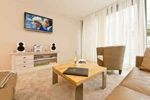 A modern living room with a wooden coffee table, a sofa, a television mounted on the wall, and a white entertainment unit with speakers.