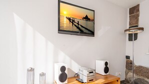 Ein modernes Wohnzimmer mit einem Holztisch, zwei Lautsprechern, einem Fernseher und einem gerahmten Sonnenuntergangsbild an der Wand.
