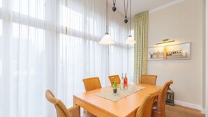 Une salle à manger avec une table en bois, des chaises en osier, des rideaux blancs et des luminaires suspendus.