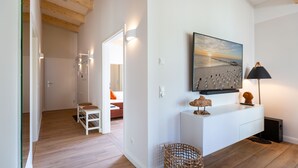 A modern living room with a wooden floor, white walls, a large painting of a beach scene, a white console table with decorative items, and a wicker basket. - Domizil Meerzeit, App. 4 (Heringsdorf)
