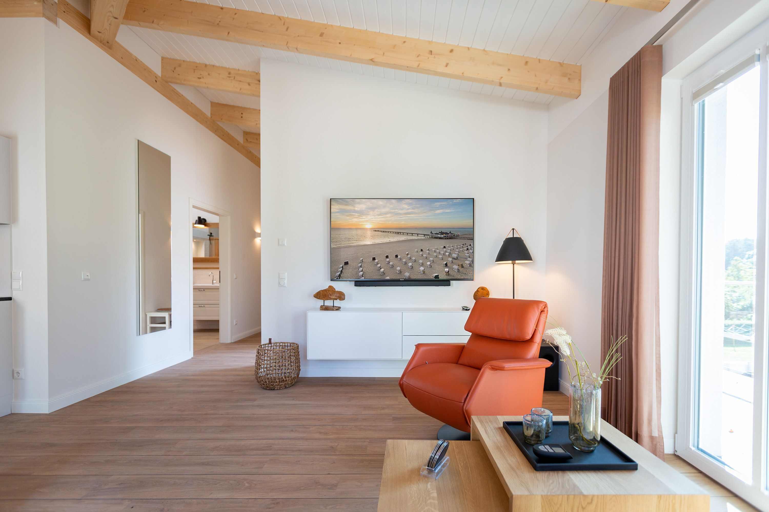 Ein modernes Wohnzimmer mit Holzboden, einem großen Fenster mit Vorhängen, einer Couch, einem Couchtisch und einem Gemälde an der Wand.