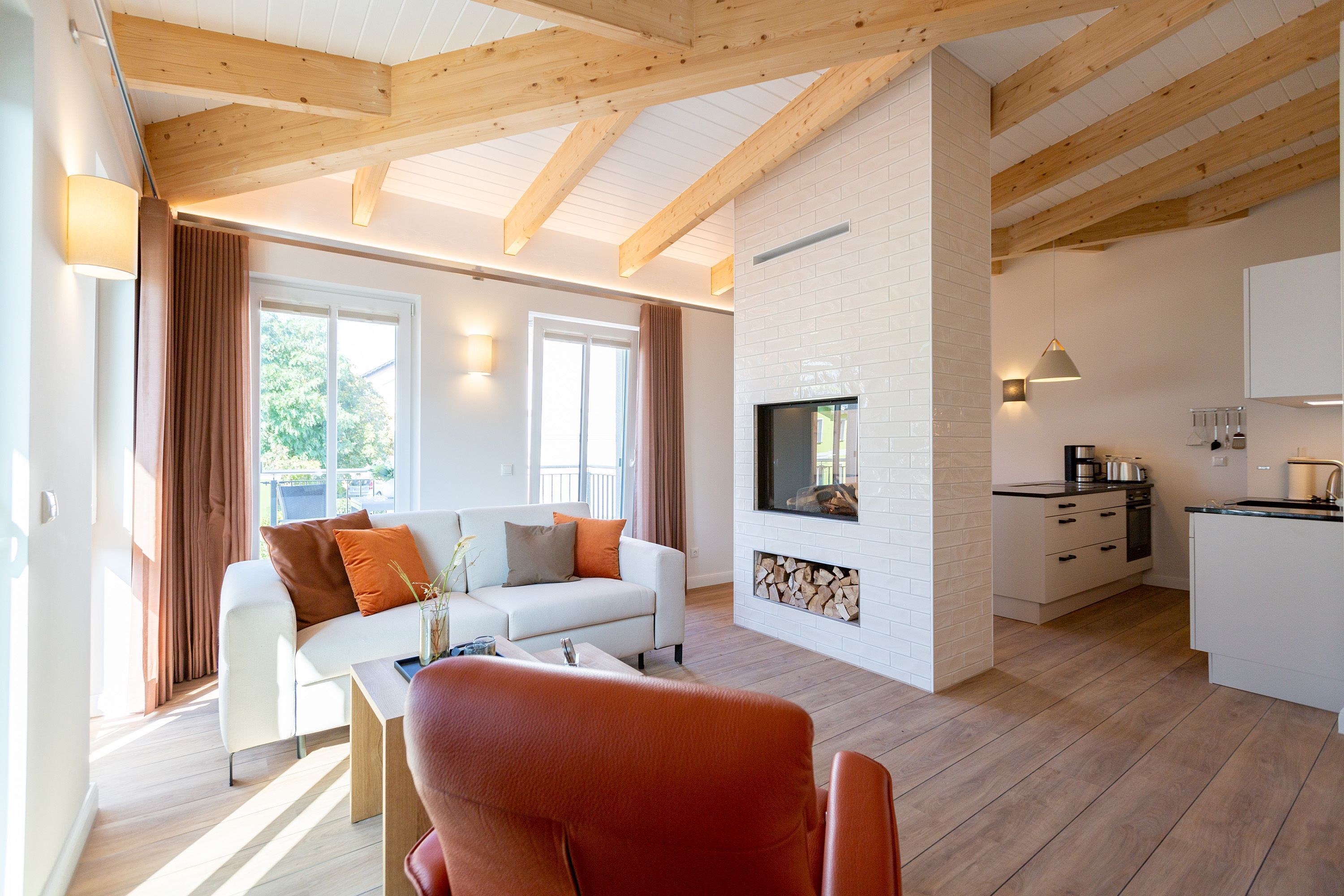 A modern living room with a white sofa, a red chair, and a fireplace. There is a kitchen area with white cabinets and a dining table.