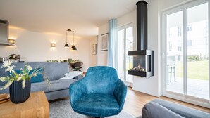 Una sala de estar moderna con un sillón azul, un sofá gris y una mesa de centro de madera. Hay una chimenea y un ventanal con vistas al exterior.