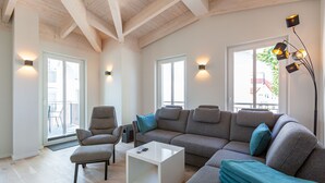 Ein modernes Wohnzimmer mit einem Ecksofa, einem weißen Couchtisch und einem grauen Sessel. Zwei Fenster bieten einen Blick nach außen, und an der Wand sind Leuchten angebracht.